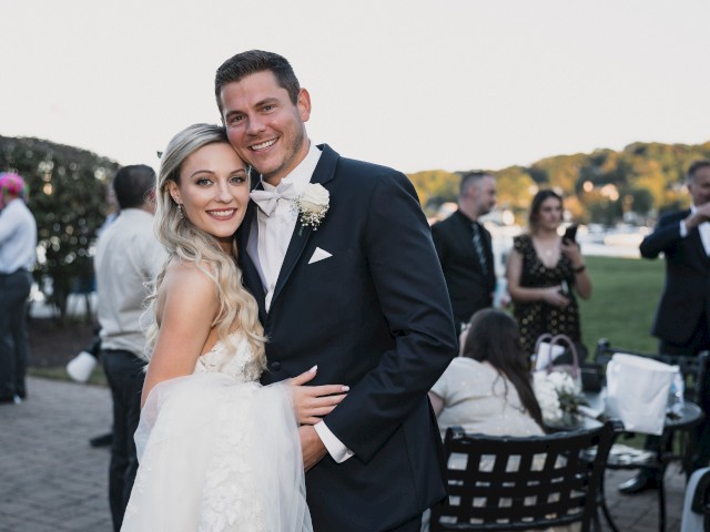 A couple, likely newlyweds, is embracing and smiling at the camera outdoors, with guests in the background at a scenic event.