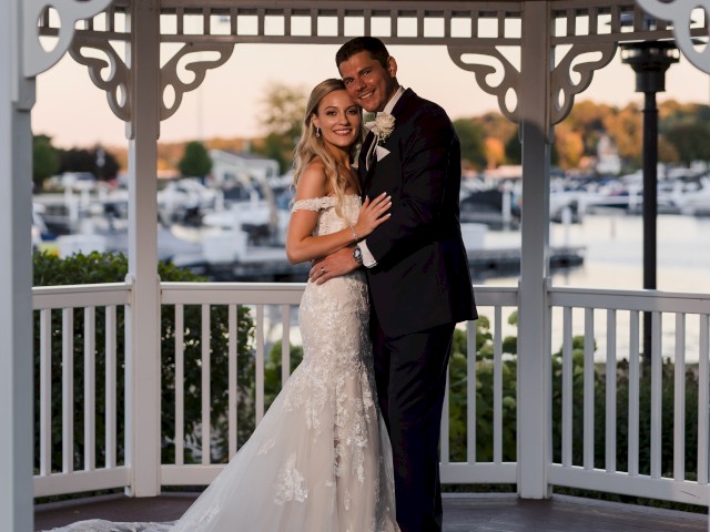 A bride and groom embrace under a gazebo with a marina in the background, both smiling and dressed in formal wedding attire.