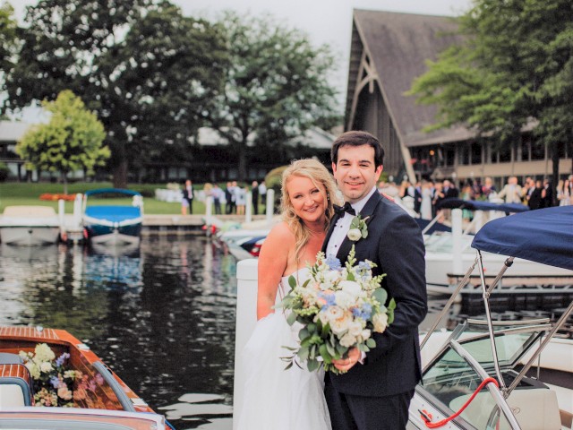 A bride and groom are standing on a dock between two boats, with the groom holding a bouquet, and a building and trees in the background, ending the sentence.