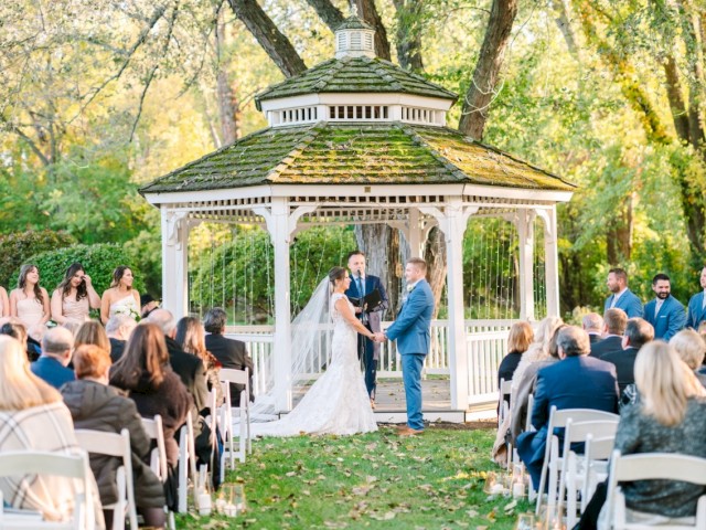 A wedding ceremony is taking place outdoors, with a couple standing in front of an officiant in a white gazebo, surrounded by guests.