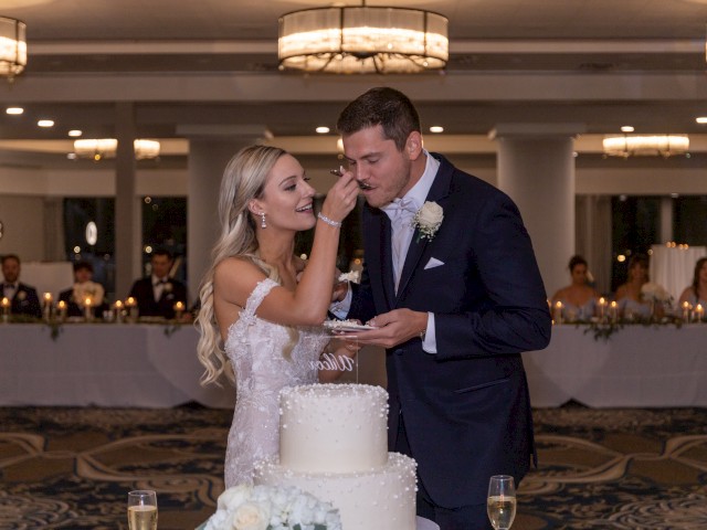 A bride feeds cake to a groom at their wedding reception, standing by a tiered cake. Both are smiling, surrounded by guests seated.