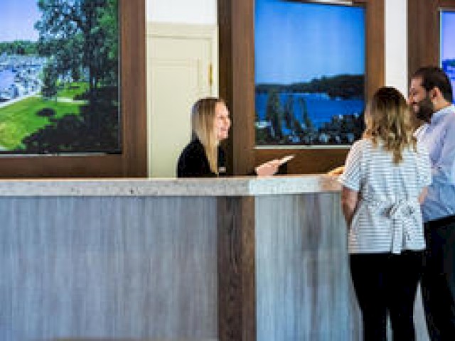 A woman behind a reception desk is assisting a couple; three landscape photos are displayed on the wall behind them.