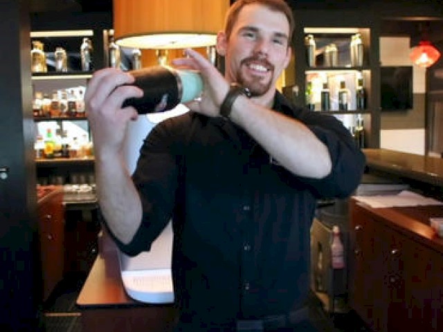 A bartender is smiling while shaking a drink shaker in a bar, with various bottles on display in the background, ending the sentence.