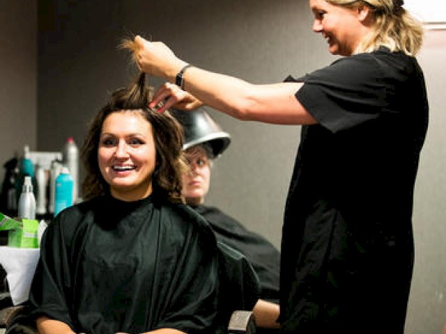A woman smiles while getting her hair done by a stylist in a salon. Another individual sits under a hair dryer in the background.