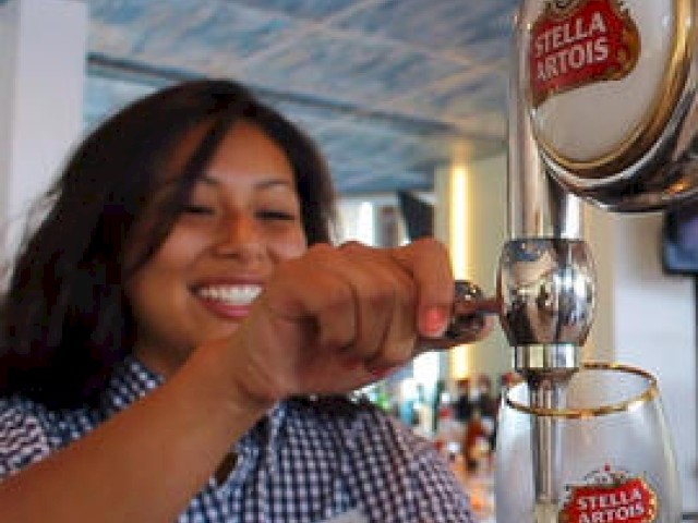 A person pours beer from a Stella Artois tap into a branded glass, smiling in a bar setting. The background is blurred.