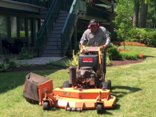 A person is mowing the lawn with a riding mower near a residential building and garden, wearing protective gear.