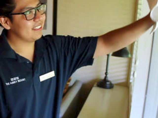 A person wearing glasses and a dark polo shirt is cleaning a window inside a room, smiling while working. A lamp and table are in the background.