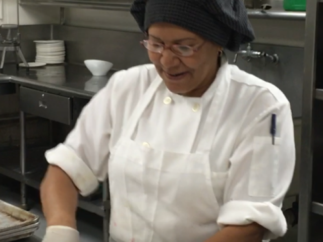 A chef in a white uniform and black hairnet is cutting dough on a counter in a commercial kitchen, with shelves and utensils in the background.