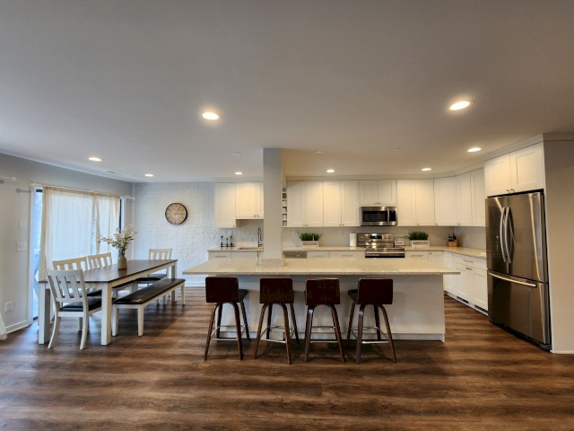A modern kitchen and dining area with white cabinets, stainless steel appliances, an island with barstools, and a dining table with chairs and benches.