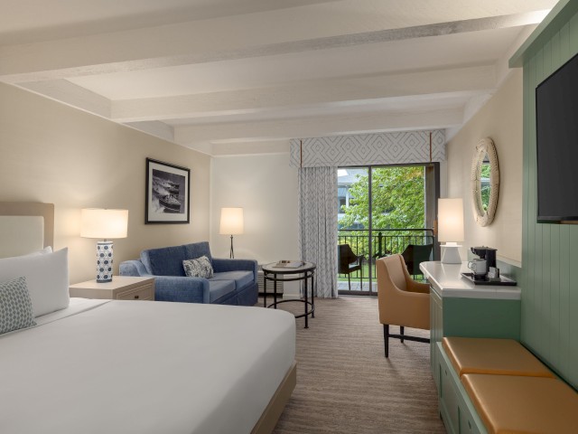 A hotel room with a bed, sofa, TV, and balcony view. There's a desk with a chair, lamps, and decor in a neutral color scheme.