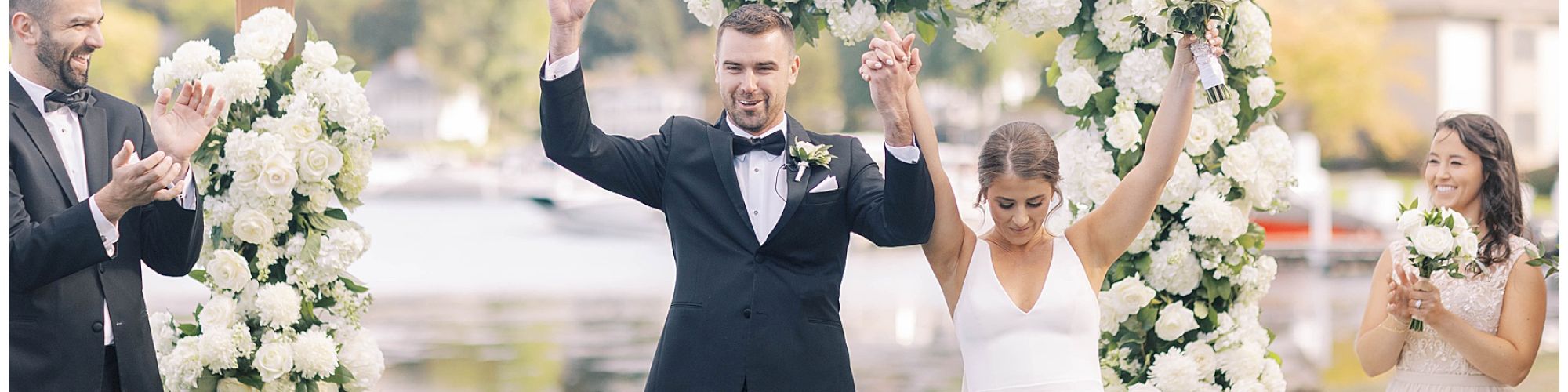 A newlywed couple celebrates under a floral arch with two people on each side applauding, situated outdoors by a lake or river.