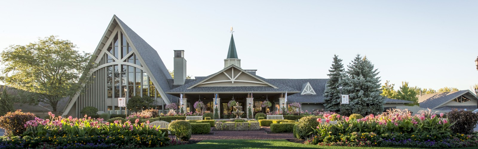 A building with a steeple and A-frame structure, surrounded by well-manicured gardens and blooming flowers under a clear sky.