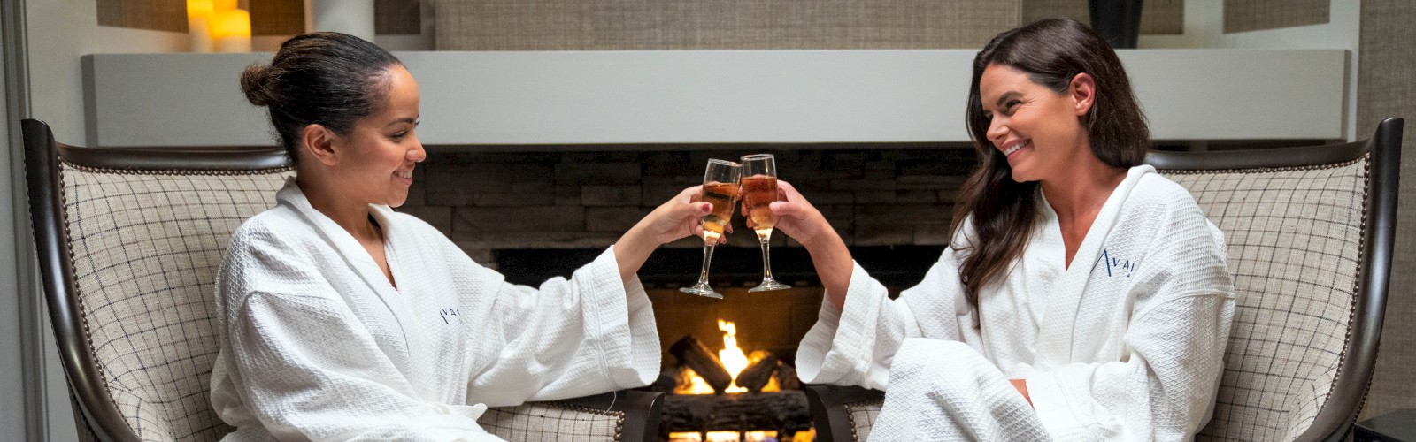 Two women in white bathrobes are sitting in chairs by a fireplace, toasting with glasses of champagne, with candles in the background.