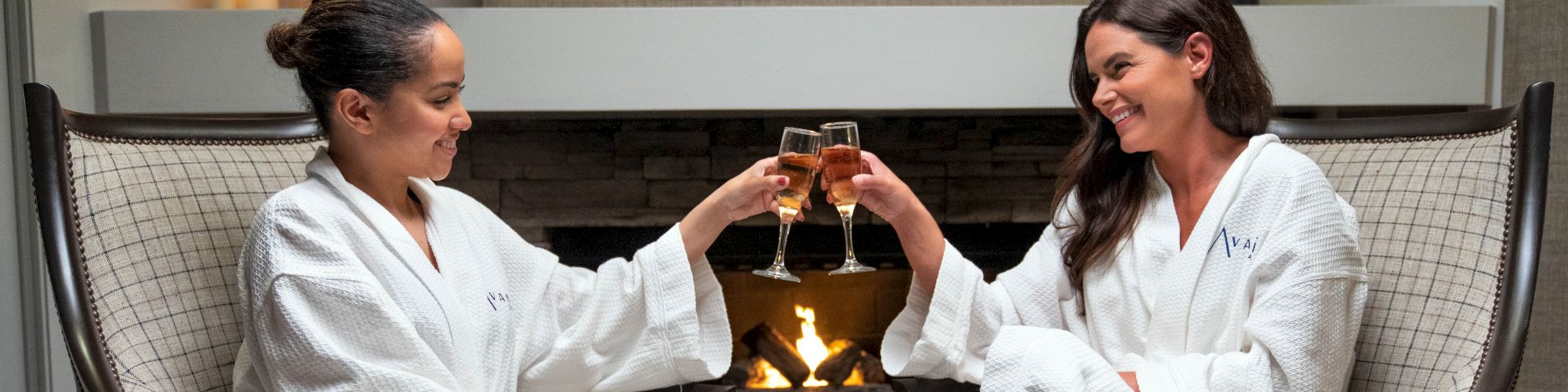 Two women in white bathrobes are sitting in chairs by a fireplace, toasting with glasses of champagne, with candles in the background.