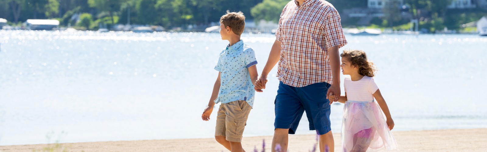 A man walks hand-in-hand with two children near a lake, with greenery and houses in the background. Flowers are in the foreground.