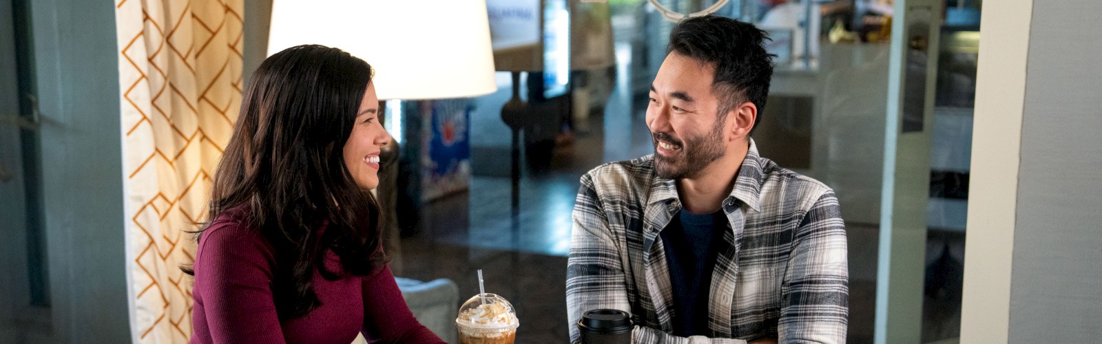 Two people sit at a wooden table in a cafe, smiling and chatting while each holds a drink. End sentence.