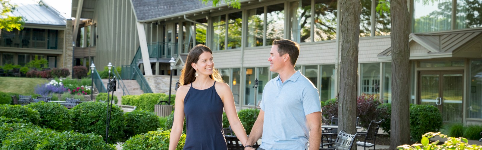 A couple is walking hand in hand on a garden pathway near a modern building with large windows and surrounded by greenery and trees.