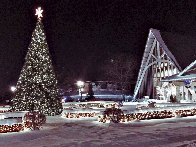 The image shows a snowy night scene with a large, decorated Christmas tree and a lit building with an A-frame roof.