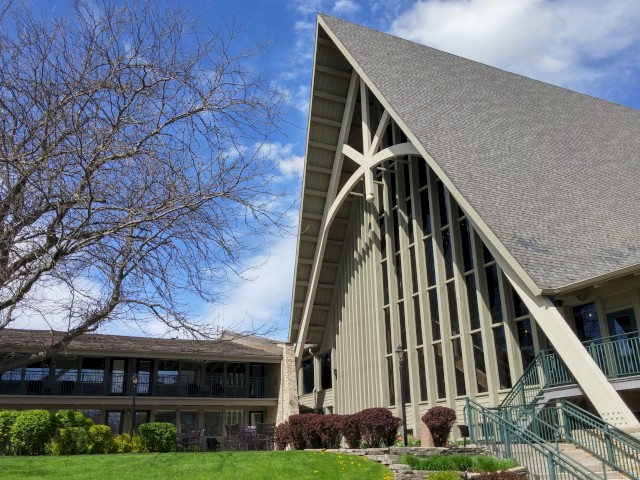 A modern building with a steep, triangular roof and large windows, accompanied by manicured greenery and a clear blue sky in the background.