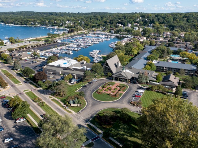 This aerial image shows a marina with numerous boats docked, surrounded by buildings, roads, and lush greenery, with a lake and wooded area in the background.