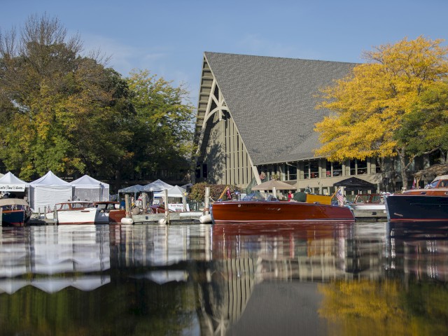 Several boats are docked near a pavilion with a large, peaked roof, surrounded by trees with autumn foliage. Tents line the waterfront.