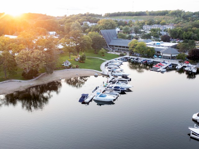 Aerial view of a marina with boats docked, a sandy beach area, trees, buildings, and a serene lake during sunset with reflections in the water.