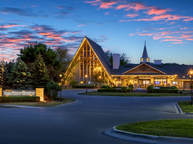 A building with triangular roof and lights is set against a twilight sky, surrounded by trees and a sign reads 