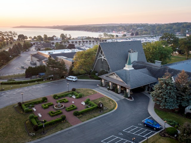 An aerial view of a large building with a landscaped garden, surrounded by trees, parking areas, and a distant waterfront during sunset.