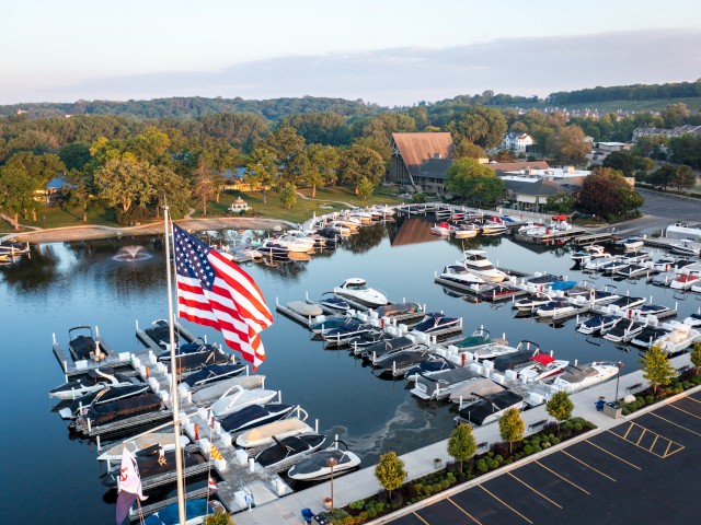A marina with numerous docked boats, an American flag, parking, and a background of trees and buildings, is by a calm river.