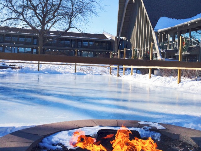 A fire pit burning in the foreground with a snowy ice rink and a modern building with string lights in the background.