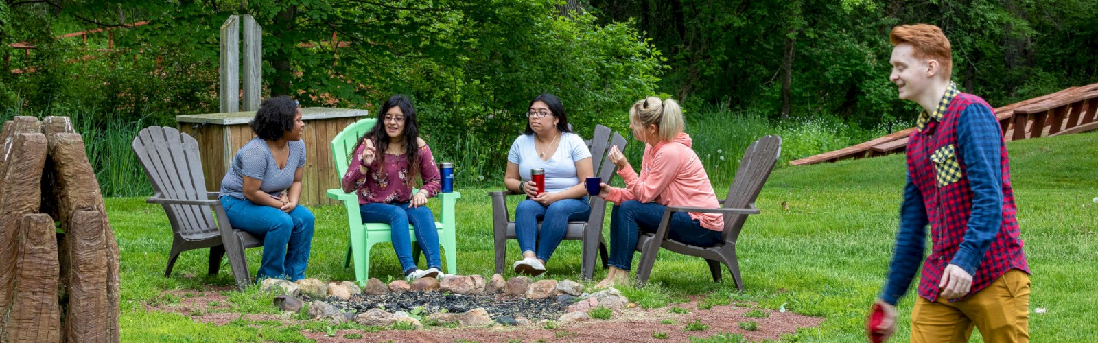 A group of people are sitting around a fire pit in a garden, while another person is playing cornhole nearby.
