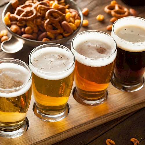 Four glasses of beer on a wooden tray with snacks like nuts and pretzels in the background on a wooden table.