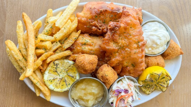 A plate of fish and chips with fries, lemon slices, sauces, coleslaw, and hush puppies on a wooden surface.