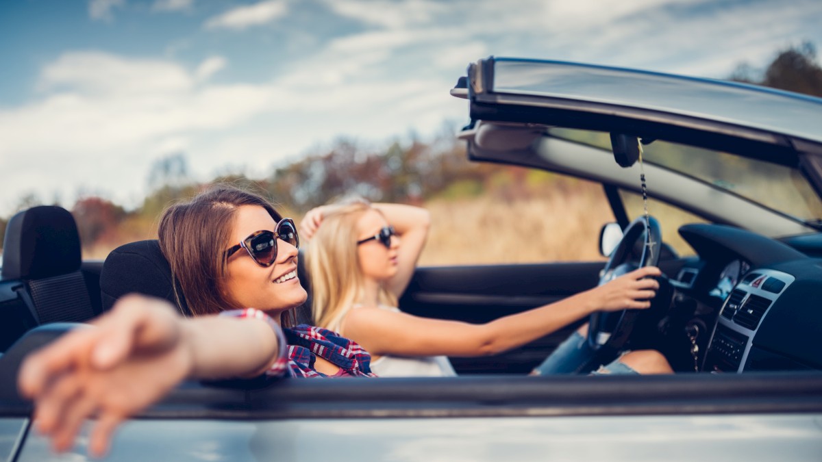 Two people in sunglasses enjoying a drive in a convertible car on a sunny day, with one person driving and the other relaxed in the passenger seat.