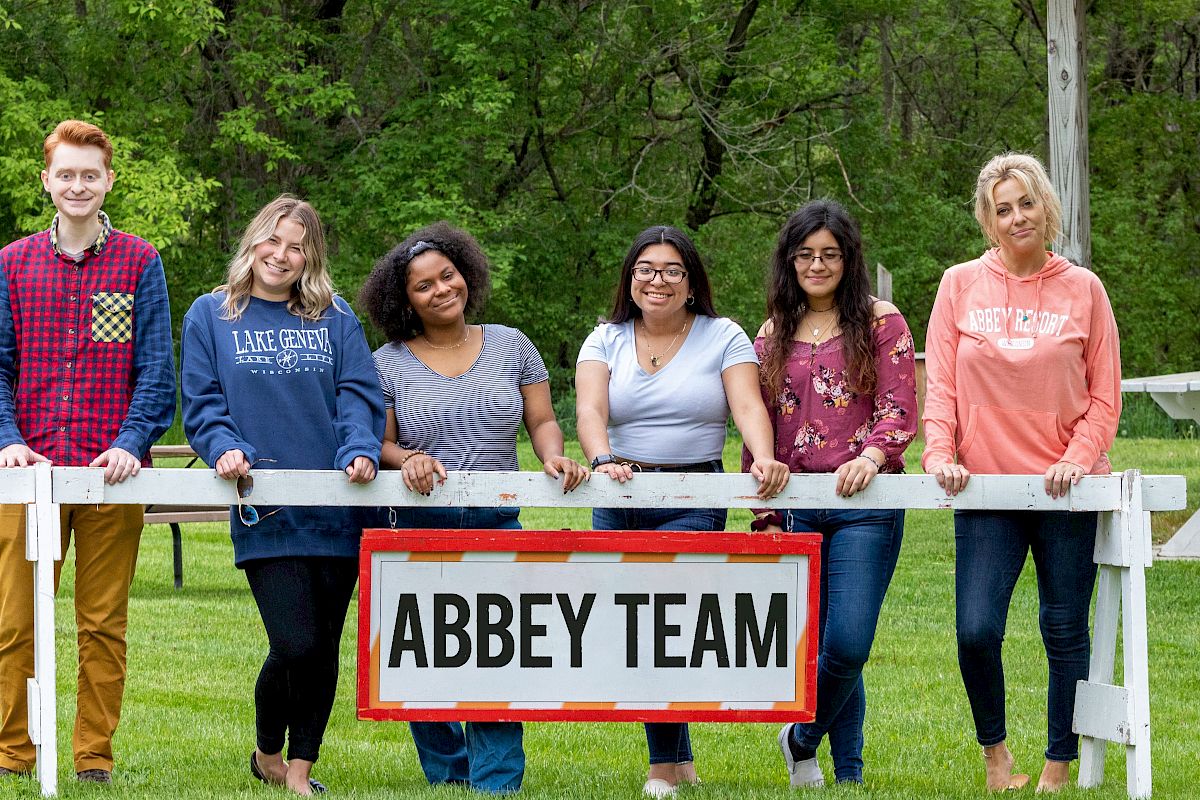 Six people are posing outdoors behind a sign that reads 