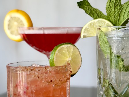 Three colorful cocktails garnished with lemon, lime, and mint, served in various glasses, are shown against a plain background.