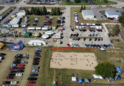 An aerial view of a fair with rides, tents, food stalls, a parking lot, and a volleyball court, surrounded by trees and buildings.
