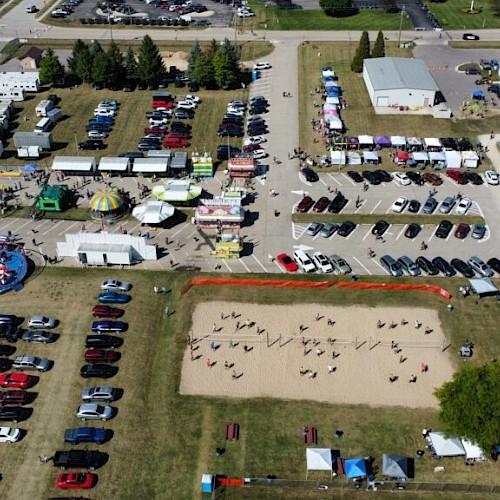 An aerial view of a fair with rides, tents, food stalls, a parking lot, and a volleyball court, surrounded by trees and buildings.