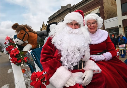 Santa and Mrs. Claus sit on a float decorated with poinsettias, with a large stuffed reindeer behind them, during a Christmas parade.