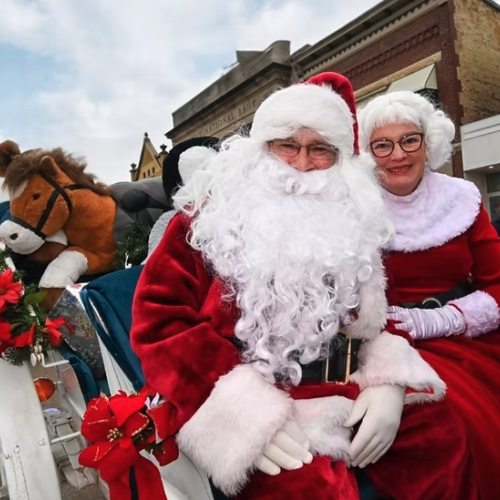 Santa and Mrs. Claus sit on a float decorated with poinsettias, with a large stuffed reindeer behind them, during a Christmas parade.