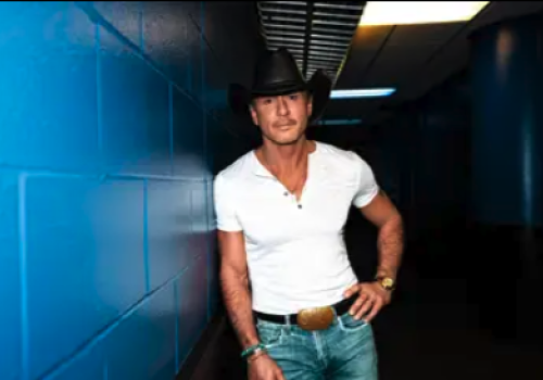 A man in a white shirt, cowboy hat, and jeans leans against a blue wall in a dimly lit corridor.