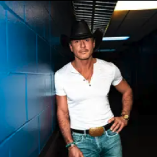 A man in a white shirt, cowboy hat, and jeans leans against a blue wall in a dimly lit corridor.