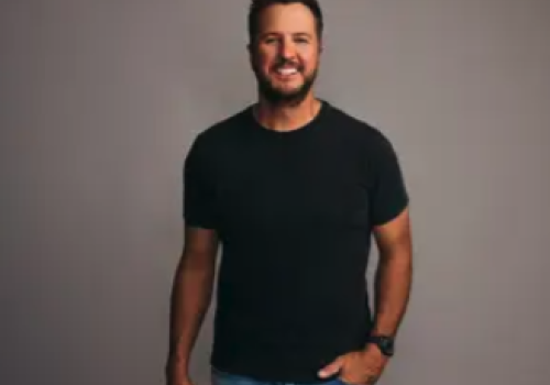 A smiling man in a black t-shirt and jeans stands against a neutral background, casually posing with one hand in his pocket.