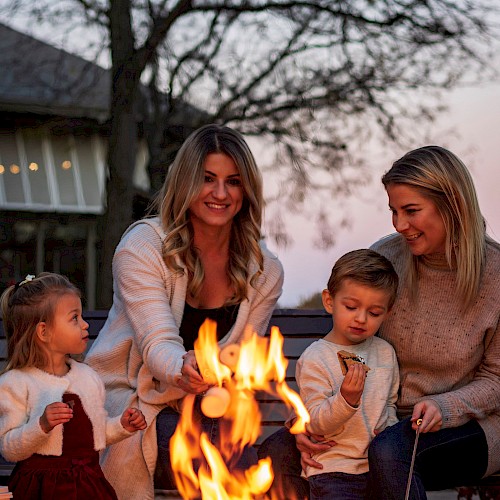 A family gathers around a firepit outdoors during sunset, enjoying snacks and smiles on a cozy evening.