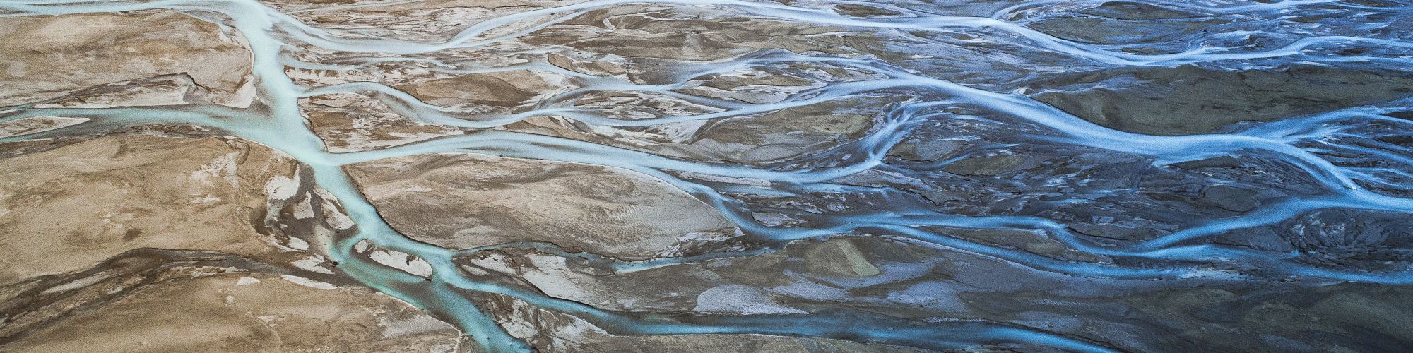 An aerial view showing a braided river flowing through a barren, rocky landscape with multiple interweaving channels of water in various shades of blue.