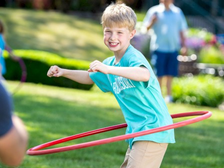 A smiling boy in a blue shirt plays with a hula hoop outside on a sunny day.