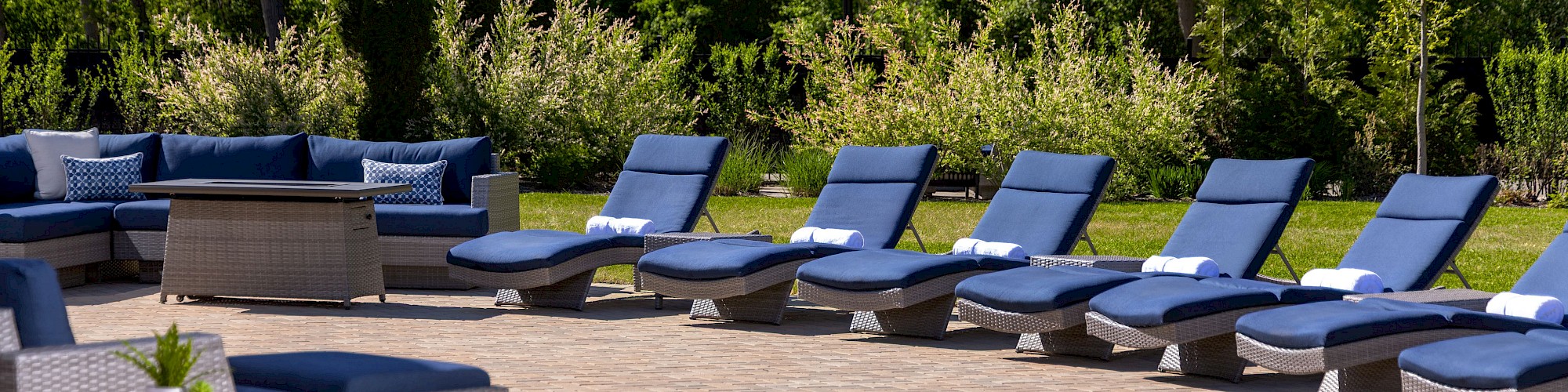 The image shows an outdoor patio area with blue lounge chairs, umbrellas, and a seating area. It's surrounded by trees and greenery.