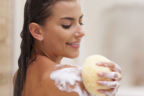 A person is in the shower, smiling while using a yellow sponge to apply soap on their shoulder, creating a lather.