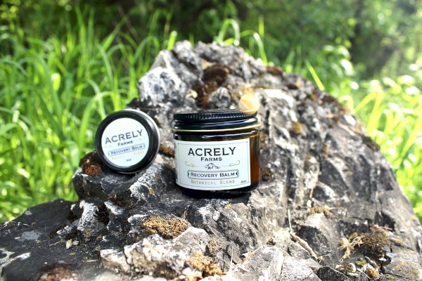 A jar and lid of Acrely Farms Recovery Balm are placed on a rock surrounded by greenery outdoors.