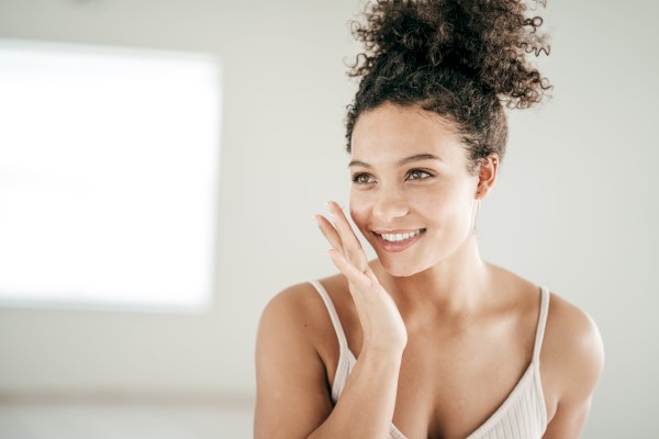 A happy woman smiles gently, touching her face, with soft natural lighting in a minimalistic, bright setting.